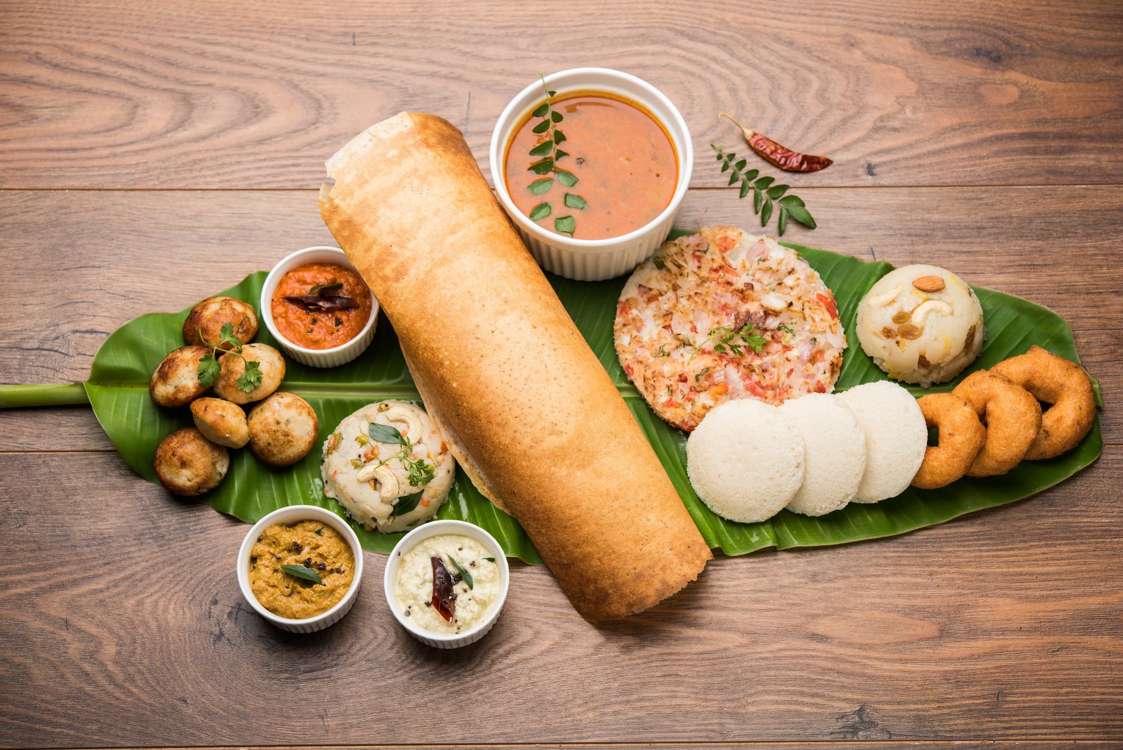 Group of South Indian food like Masala Dosa, Uttapam, Idli/idly, Wada/vada, sambar, appam, semolina halwa, upma served over banana leaf with colourful chutneys, selective focus